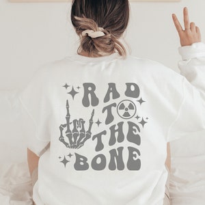 Peut inclure: Sweat-shirt blanc avec un imprim&eacute; graphique gris au dos qui dit "Rad to the Bone" avec une main de squelette faisant un doigt d'honneur et un symbole radioactif.