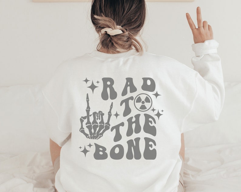 Peut inclure: Sweat-shirt blanc avec un imprim&eacute; graphique gris au dos qui dit "Rad to the Bone" avec une main de squelette faisant un doigt d'honneur et un symbole radioactif.