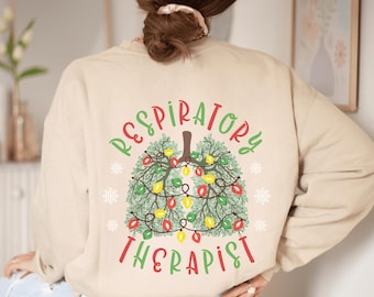 May include: Beige sweatshirt with a Christmas tree design shaped like lungs. The design includes green, red, and yellow colors, with the words "Respiratory Therapist" in a festive font. Snowflakes are also included in the design.