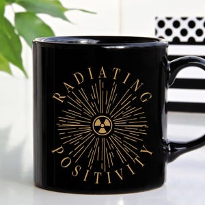 May include: Black ceramic mug with the words "RADIATING POSITIVITY" in gold lettering. The design includes a sunburst around a radiation symbol. The mug has a curved handle and is set against a neutral background.