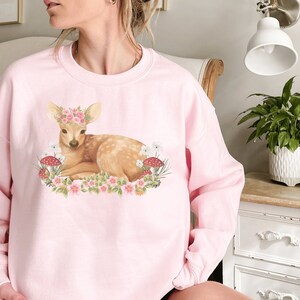 May include: A light pink sweatshirt featuring a watercolor illustration of a deer wearing a floral crown, surrounded by flowers and mushrooms. The deer is lying down. The sweatshirt is a crew neck style.