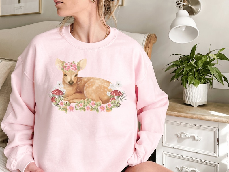 May include: A light pink sweatshirt featuring a watercolor illustration of a deer wearing a floral crown, surrounded by flowers and mushrooms. The deer is lying down. The sweatshirt is a crew neck style.