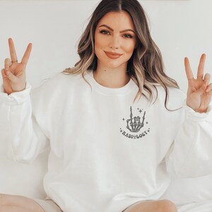 Peut inclure: Sweat-shirt blanc avec une main de squelette grise faisant un signe de rock and roll avec le texte "RADIOLOGY" en dessous.