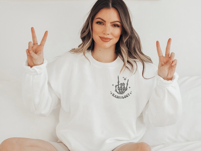 Peut inclure: Sweat-shirt blanc avec une main de squelette grise faisant un signe de rock and roll avec le texte "RADIOLOGY" en dessous.