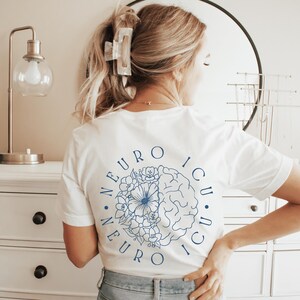 May include: White t-shirt with a blue floral and brain design, and the text "NEURO ICU" in a circular pattern. The shirt is worn by a person with blonde hair, and is paired with blue jeans.