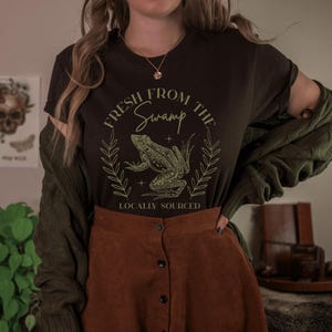 May include: A dark brown t-shirt with the text "Fresh From The Swamp" and a frog graphic. The design is in a light green color and includes the words "Locally Sourced". The shirt is paired with a brown skirt and a green cardigan.