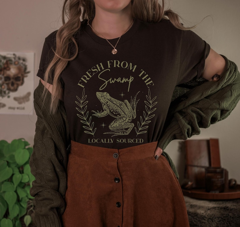 May include: A dark brown t-shirt with the text "Fresh From The Swamp" and a frog graphic. The design is in a light green color and includes the words "Locally Sourced". The shirt is paired with a brown skirt and a green cardigan.