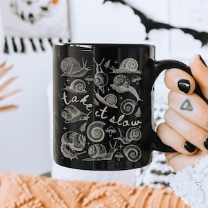 May include: Black ceramic mug featuring a detailed illustration of various snails and mushrooms. The mug has the phrase "take it slow" in a handwritten style. The mug is held by a person with black nail polish.