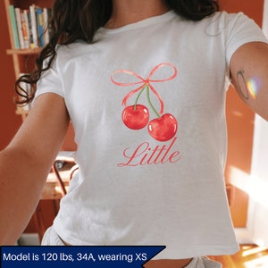 May include: White t-shirt with a red cherry graphic and the word "Little" printed in pink. The model is wearing a size XS.