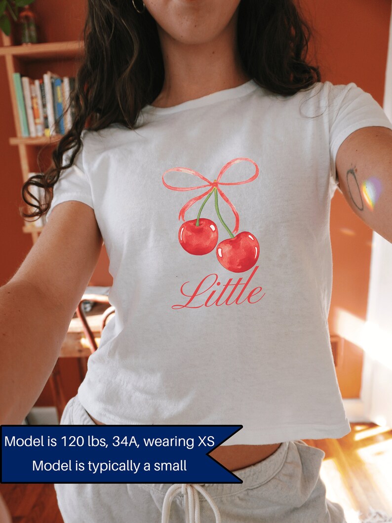 May include: White t-shirt with a red cherry graphic and the word "Little" printed in pink. The model is wearing a size XS.
