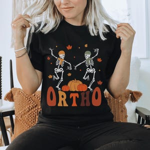 Könnte beinhalten: Schwarzes T-Shirt mit einem Motiv von zwei Skeletten, die auf Kürbissen tanzen, mit dem Text "ORTHO" in orangefarbenen und gelben Buchstaben.