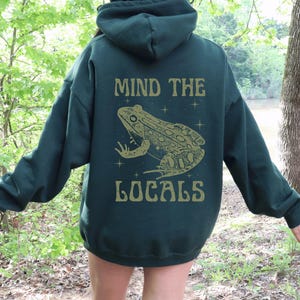 Può includere: Una felpa con cappuccio verde scuro con la scritta "MIND THE LOCALS" e una grafica di rana dorata sul retro. La felpa ha un cappuccio e maniche lunghe. L'immagine è scattata all'aperto.
