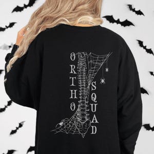 Könnte beinhalten: Schwarzes Sweatshirt mit weißem Grafikdesign, das eine Wirbelsäule, Spinnweben und Spinnen zeigt. Das Wort "ORTHOSQUAD" ist vertikal auf der Rückseite aufgedruckt. Das Sweatshirt ist mit schwarzen Fledermaus-Ausschnitten verziert.