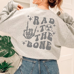 Peut inclure: Sweat-shirt gris avec un imprim&eacute; graphique d'une main de squelette faisant un doigt d'honneur, un symbole nucl&eacute;aire et le texte "Rad to the Bone".