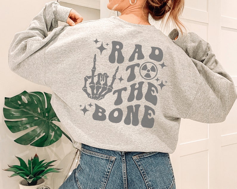 Peut inclure: Sweat-shirt gris avec un imprim&eacute; graphique d'une main de squelette faisant un doigt d'honneur, un symbole nucl&eacute;aire et le texte "Rad to the Bone".