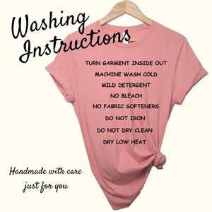 Pu&ograve; includere: Una maglietta rosa con testo nero che recita "Washing Instructions" e elenca le istruzioni di cura per il capo. Il testo include: Girare il capo al rovescio, Lavaggio in lavatrice a freddo, Detersivo delicato, Non candeggiare, Non usare ammorbidente, Non stirare, Non lavare a secco, Asciugare a bassa temperatura. La parte inferiore della maglietta recita "Handmade with care just for you".