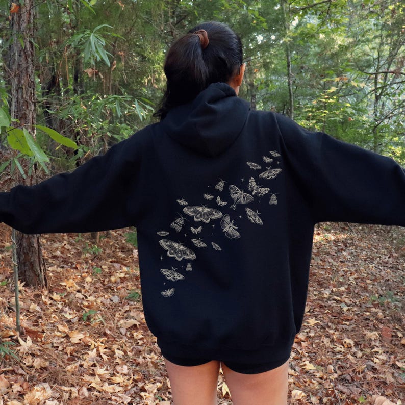 May include: Black hoodie with a beige moth and butterfly design on the back. The design features various sizes and shapes of moths and butterflies. The hoodie has a hood and long sleeves. The person is standing outside in a forest.