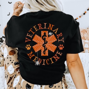 Puede incluir: Camiseta negra con un símbolo de medicina veterinaria naranja y negro con temática de Halloween. El símbolo incluye un caduceo, una telaraña y huellas de patas. El texto "Medicina Veterinaria" está escrito alrededor del símbolo.