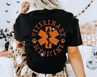 Peut inclure: T-shirt noir avec un symbole de médecine vétérinaire orange et noir sur le thème d'Halloween. Le symbole comprend un caducée, une toile d'araignée et des empreintes de pattes. Le texte "Médecine vétérinaire" est écrit autour du symbole.