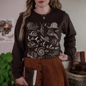 Puede incluir: Una sudadera marr&oacute;n oscuro con una ilustraci&oacute;n de caracoles y setas de color crema. El texto "take it slow" est&aacute; escrito en cursiva. La sudadera se combina con una falda marr&oacute;n.