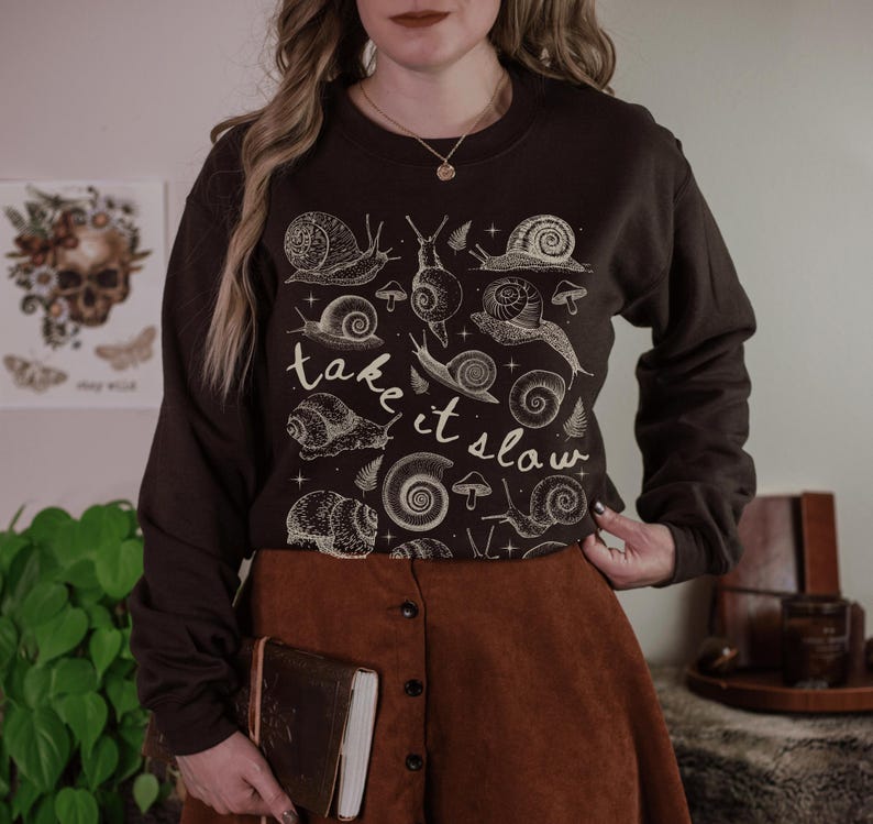Puede incluir: Una sudadera marr&oacute;n oscuro con una ilustraci&oacute;n de caracoles y setas de color crema. El texto "take it slow" est&aacute; escrito en cursiva. La sudadera se combina con una falda marr&oacute;n.