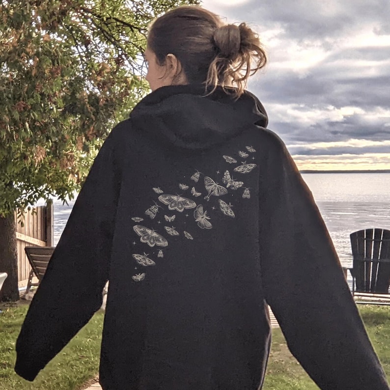 May include: Black hoodie with a hood and long sleeves. The back of the hoodie features a design of several moths in various sizes and shapes. The moths are in a light color, contrasting with the dark fabric of the hoodie.