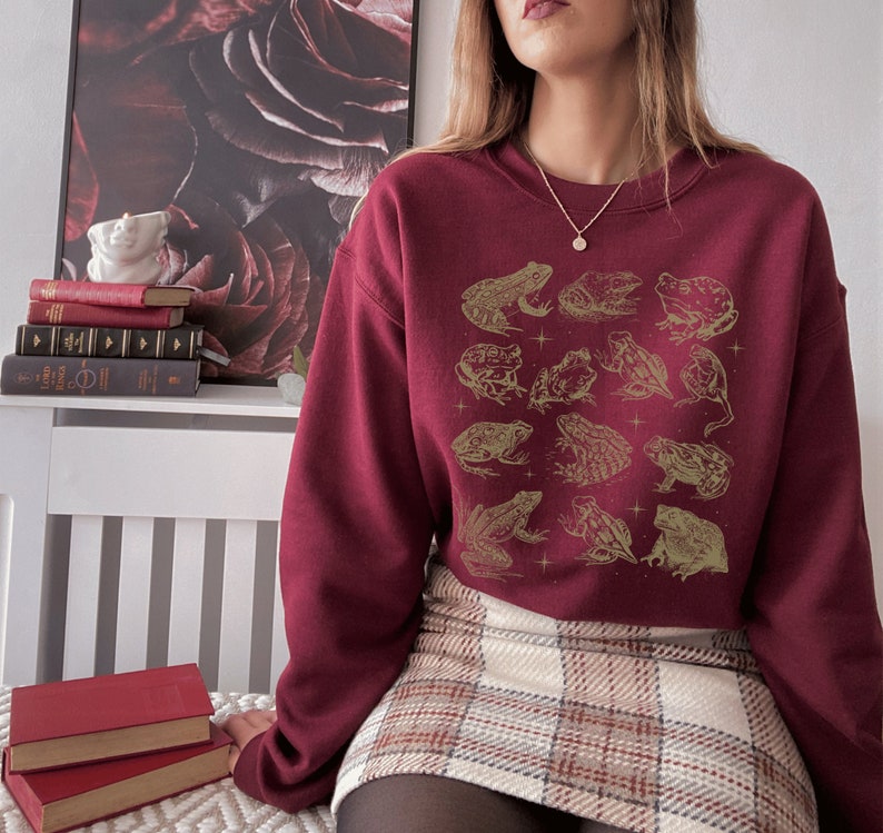 May include: A maroon sweatshirt with a graphic print of frogs in gold. The sweatshirt has a crew neck and long sleeves.