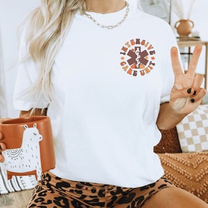 Peut inclure: T-shirt blanc avec un motif circulaire indiquant "Intensive Care Unit" en marron et orange. Le motif comprend un symbole m&eacute;dical, des citrouilles et des feuilles. La personne porte un short &agrave; imprim&eacute; l&eacute;opard et tient une tasse lama marron.