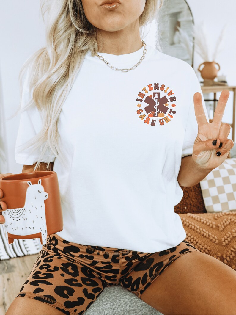 Peut inclure: T-shirt blanc avec un motif circulaire indiquant "Intensive Care Unit" en marron et orange. Le motif comprend un symbole m&eacute;dical, des citrouilles et des feuilles. La personne porte un short &agrave; imprim&eacute; l&eacute;opard et tient une tasse lama marron.