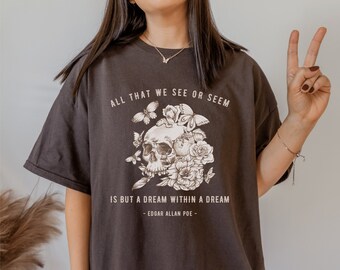 May include: A dark gray t-shirt with a graphic print of a skull surrounded by flowers and butterflies. The text "ALL THAT WE SEE OR SEEM IS BUT A DREAM WITHIN A DREAM - EDGAR ALLAN POE -" is printed on the shirt.