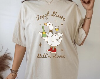 Puede incluir: Camiseta beige con un diseño de ganso de dibujos animados. El ganso sostiene una botella de champán y una copa de vino. Lleva pajarita y zapatos rojos. El texto en la camiseta dice "Legal Goose" y "Gett'n Loose."