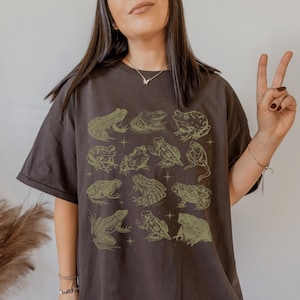 Pode incluir: Uma camiseta marrom escura com uma estampa gr&aacute;fica de sapos em v&aacute;rias poses. Os sapos s&atilde;o delineados em uma cor verde clara e est&atilde;o dispostos em uma grade.