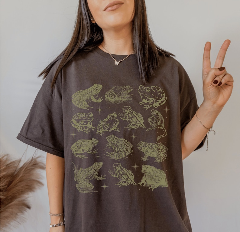 Pode incluir: Uma camiseta marrom escura com uma estampa gr&aacute;fica de sapos em v&aacute;rias poses. Os sapos s&atilde;o delineados em uma cor verde clara e est&atilde;o dispostos em uma grade.