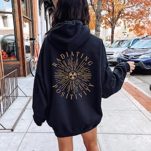 Może przedstawiać: Czarna bluza z kapturem z napisem "RADIATING POSITIVITY" w złotym wzorze promieni słonecznych na plecach. Projekt zawiera symbol promieniowania. Noszona na zewnątrz.