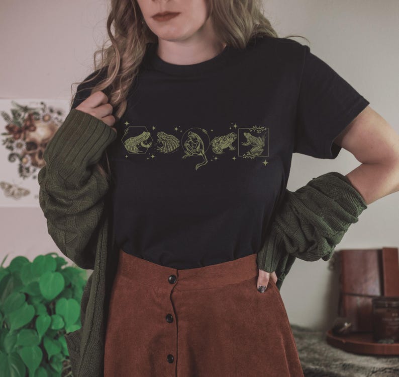 Pu&ograve; includere: Una maglietta nera con una linea di cinque rane in un design circolare. Le rane sono contornate d'oro e riempite di verde. La maglietta &egrave; indossata da una persona con i capelli lunghi castani e una gonna marrone.