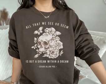 Op de afbeelding: Een bruin sweatshirt met een schedel- en bloemenontwerp. De tekst "ALL THAT WE SEE OR SEEM IS BUT A DREAM WITHIN A DREAM - EDGAR ALLAN POE -" staat op de voorkant gedrukt.