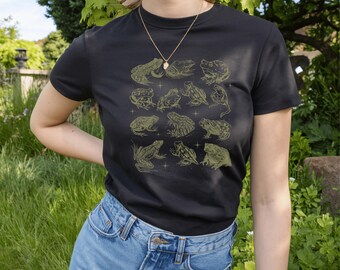 May include: Black t-shirt featuring a collection of green frog illustrations. The frogs are in various poses and sizes, with a few small star-like accents. The shirt is paired with blue denim shorts.