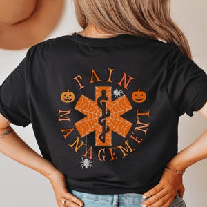 May include: Black t-shirt with a Halloween-themed design featuring a medical symbol with a spider web, pumpkins, and the text "Pain Management".