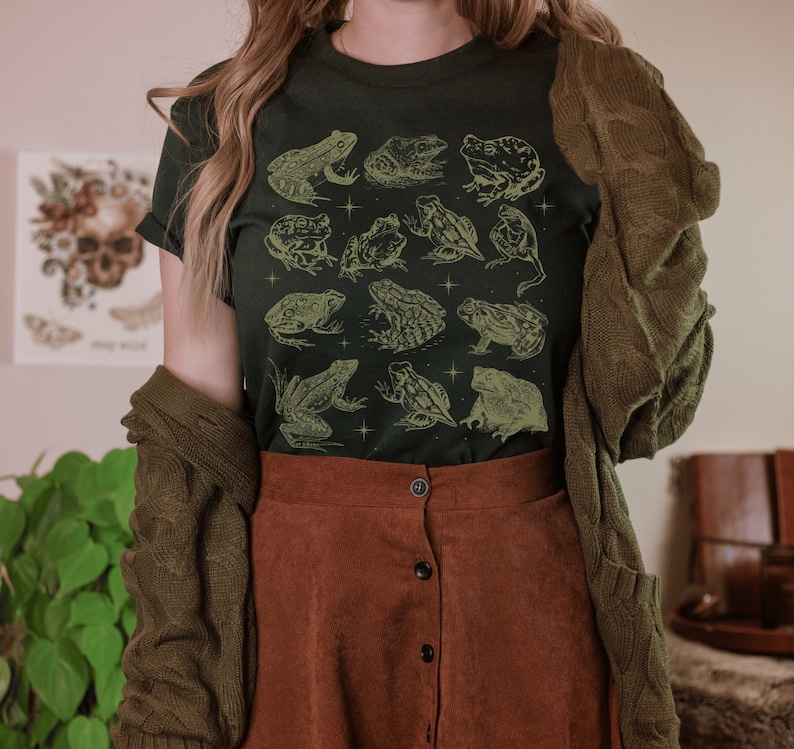Pode incluir: Uma camiseta verde escura com um padr&atilde;o de sapos em v&aacute;rias poses. Os sapos s&atilde;o delineados em uma cor verde clara e t&ecirc;m uma est&eacute;tica vintage.