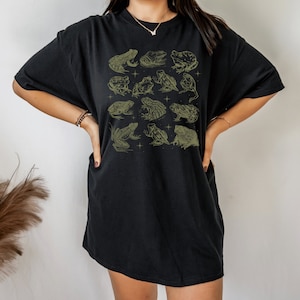 Pode incluir: Camiseta preta oversized com uma estampa gr&aacute;fica de v&aacute;rios sapos e r&atilde;s em verde claro. A camiseta tem um caimento solto e um decote redondo.