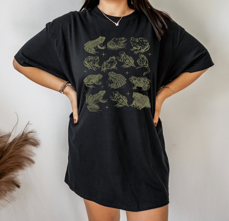 Pode incluir: Camiseta preta oversized com uma estampa gr&aacute;fica de v&aacute;rios sapos e r&atilde;s em verde claro. A camiseta tem um caimento solto e um decote redondo.