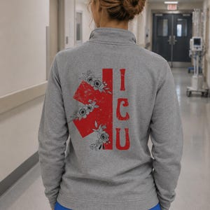 Peut inclure: Un pull gris chiné à fermeture éclair au col, orné au dos d'une étoile de vie médicale rouge à l'aspect vieilli. Le symbole est décoré d'illustrations florales en traits noirs. Les lettres ICU sont imprimées en rouge à côté du symbole.