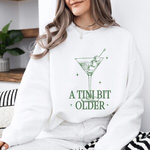 May include: White sweatshirt with a green martini glass graphic and the text "A TINI BIT OLDER".