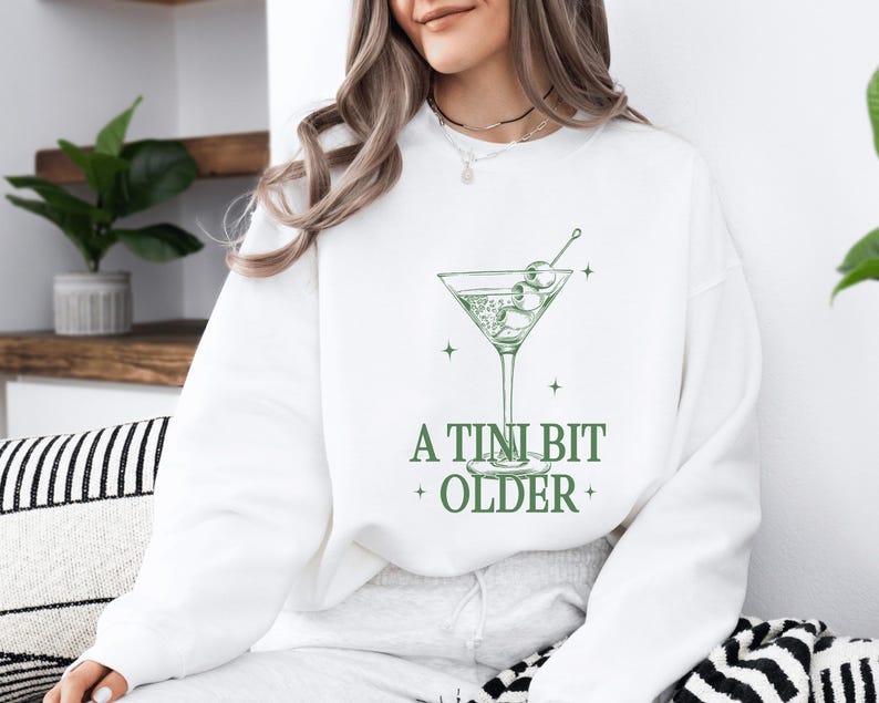 May include: White sweatshirt with a green martini glass graphic and the text "A TINI BIT OLDER".