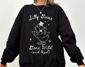 Puede incluir: Sudadera negra con un gráfico blanco de un ganso con sombrero de copa, sosteniendo una botella y una copa de vino. El texto dice "Silly Goose Gone Wild (and legal)". La sudadera tiene mangas largas y cuello redondo.