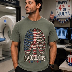 May include: A sage green crew neck t-shirt featuring a graphic of a human rib cage filled with a red, white, and blue American flag pattern. Below the rib cage, the word RADIOLOGY is printed in white, flanked by small red and blue stars.