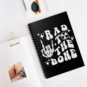 Könnte beinhalten: Schwarzer Spiralblock mit einem weißen Design, das "Rad to the Bone" sagt, mit einer Skeletthand, die das Rock'n'Roll-Zeichen macht.