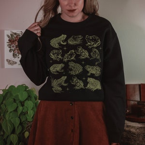 May include: Black sweatshirt with a green and white design of various frogs. The sweatshirt has a crew neck and long sleeves.