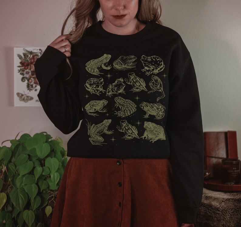 May include: Black sweatshirt with a green and white design of various frogs. The sweatshirt has a crew neck and long sleeves.