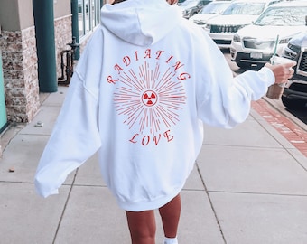 May include: White hoodie with a red graphic design on the back. The design features the words "RADIATING LOVE" around a sunburst with a radiation symbol. The hoodie has a hood and long sleeves. The person is walking outside.